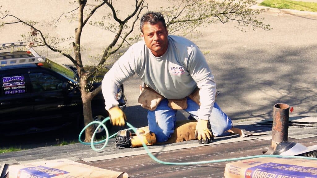 Man on a roof fastening roof shingles with a nail gun.
